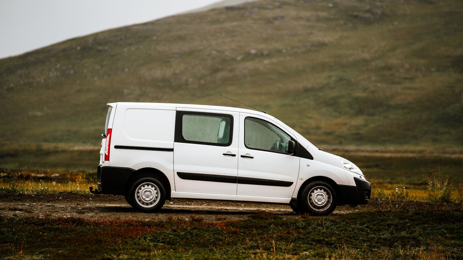 A white van parked on a rural road with a scenic mountain backdrop under a cloudy sky.